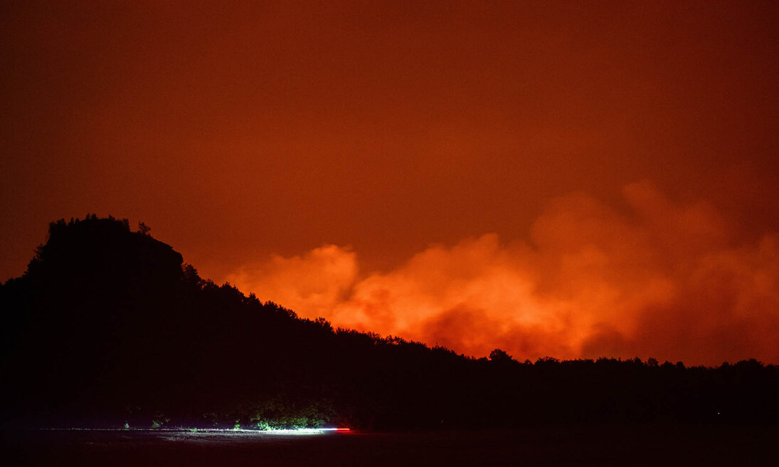 Qualm zieht in einer Nacht hinter einem Berg während eines Waldbrandes auf. Die Nacht wird durch das Feuer erleuchtet.
