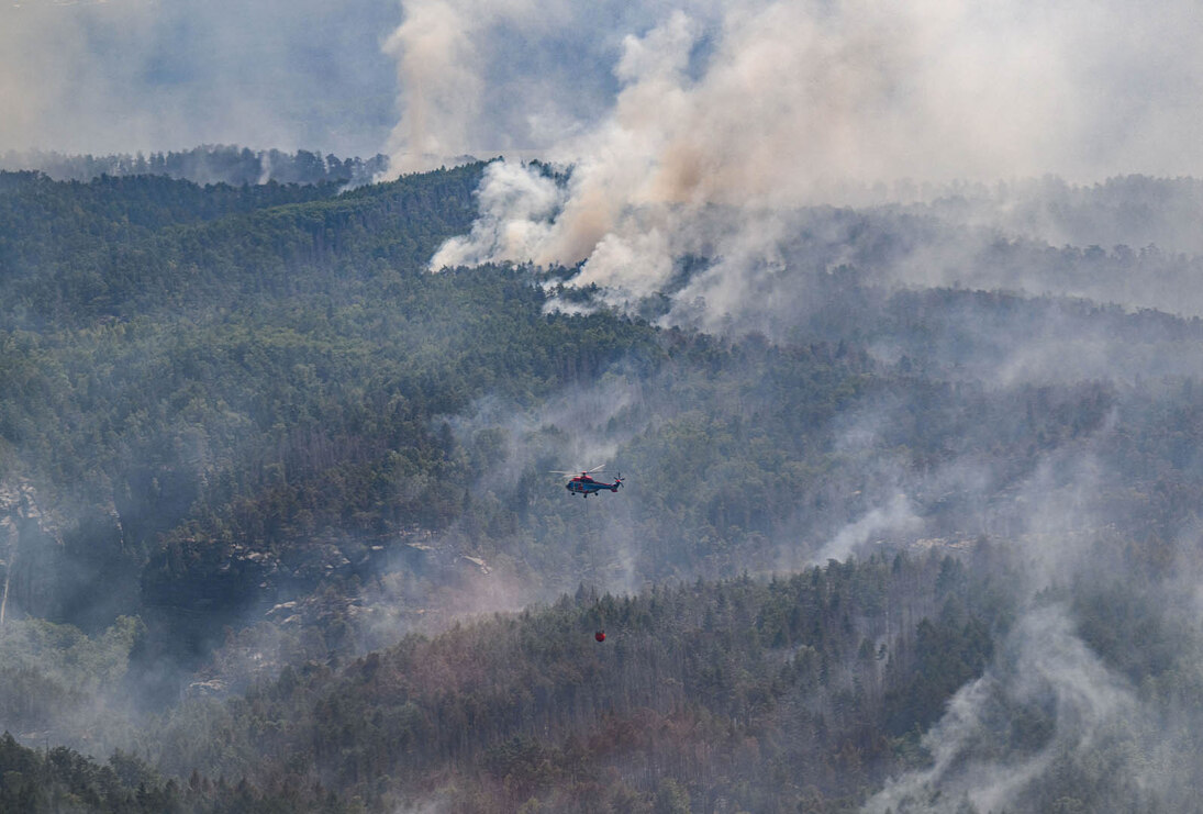 Ein Helikopter fliegt mit einem Wasserbehälter über einen Wald. Aus dem Wald sind vereinzelt weiße Rauchschwaden zu sehen.