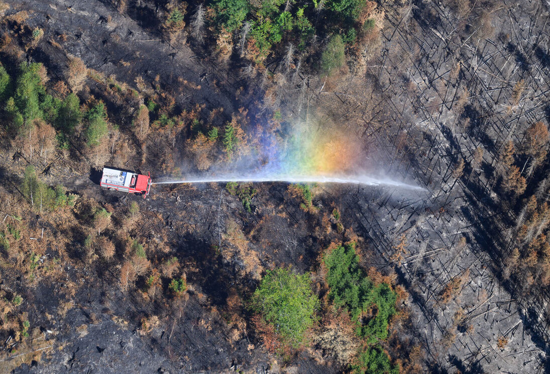 Ein Feuerwehrauto wird von oben fotografiert. Im Strahl des Löschwassers einer Feuerwehr bildet sich bei den Löscharbeiten der Waldbrände im Nationalpark Sächsische Schweiz ein Regenbogen.