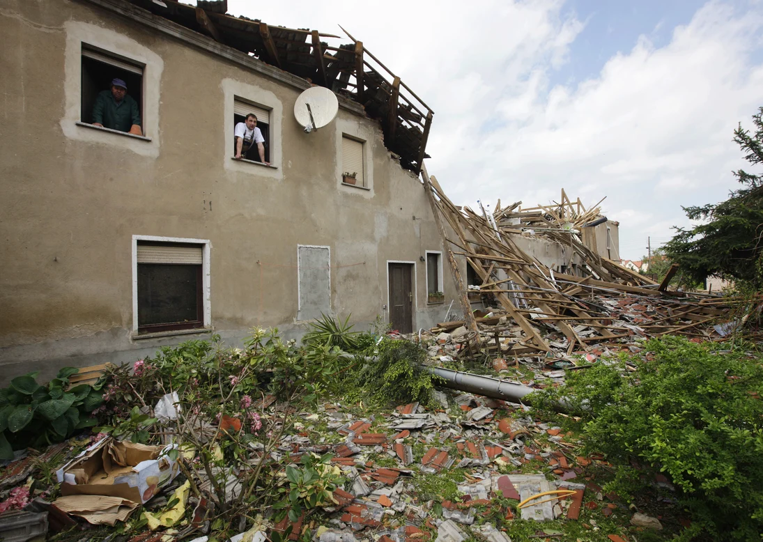 Ein nahezu vollständig durch Sturm zerstörtes Haus, aus dessen Fenstern im Obergeschoss zwei Männer schauen.