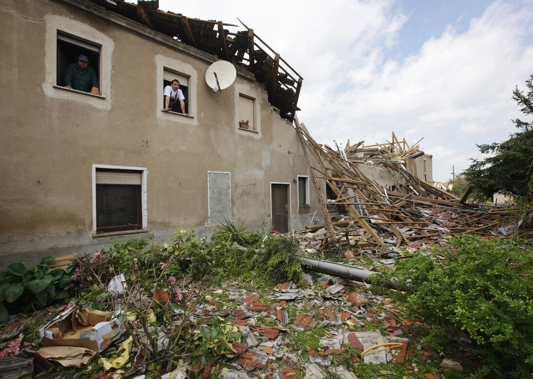 Ein nahezu vollständig durch Sturm zerstörtes Haus, aus dessen Fenstern im Obergeschoss zwei Männer schauen.